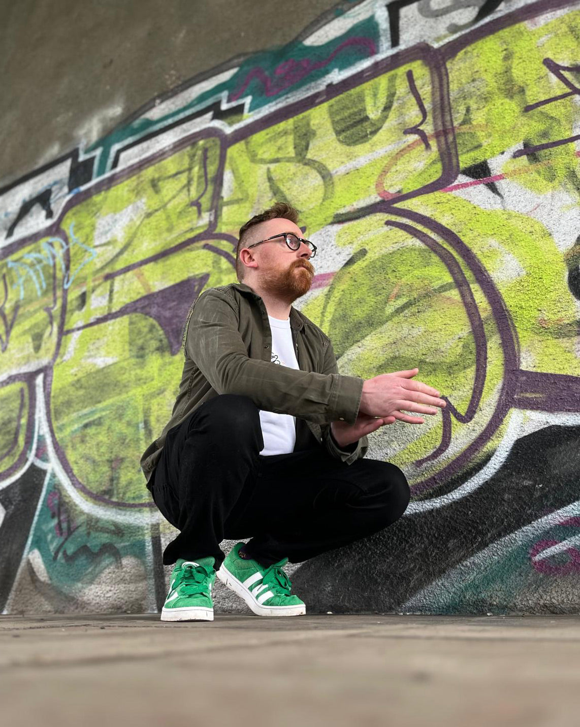 Artist Mark Gilmour in front of a graffiti wall wearing green Adidas sneakers.